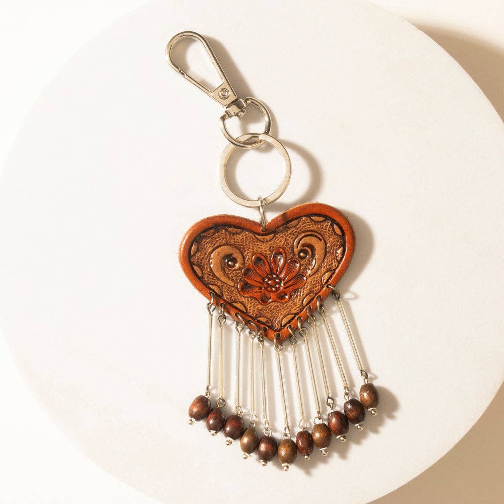A heart-shaped brown leather keychain with hand-tooled floral details, silver-tone metal rings, and dangling silver chains ending in small brown beads, displayed on a white circular surface.