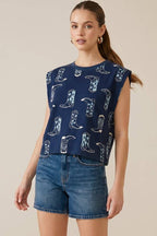 Woman wearing a navy sleeveless top with cowboy boot pattern and denim shorts on a beige background