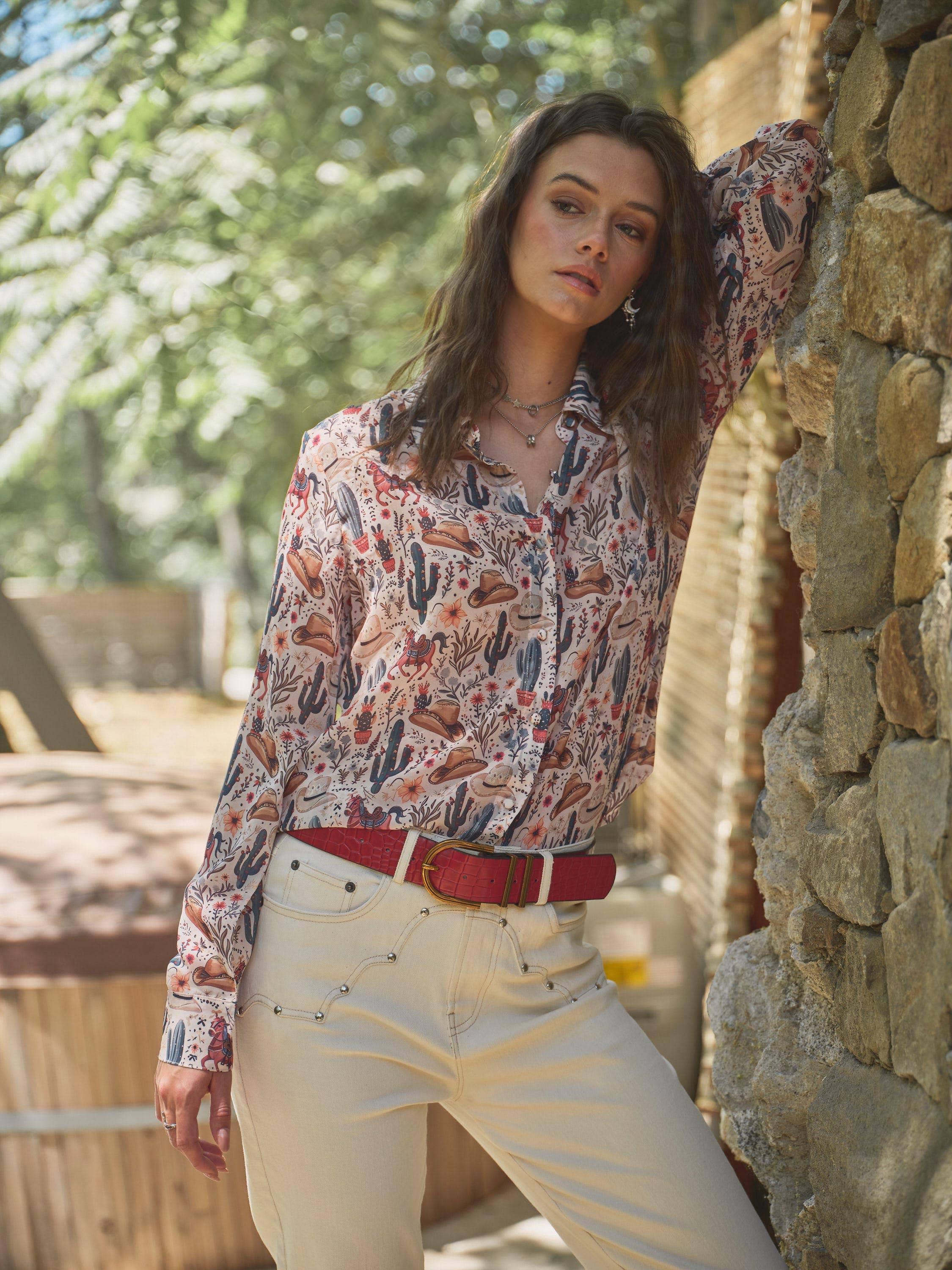 Woman wearing a patterned shirt and beige pants leaning against a stone wall outdoors.
