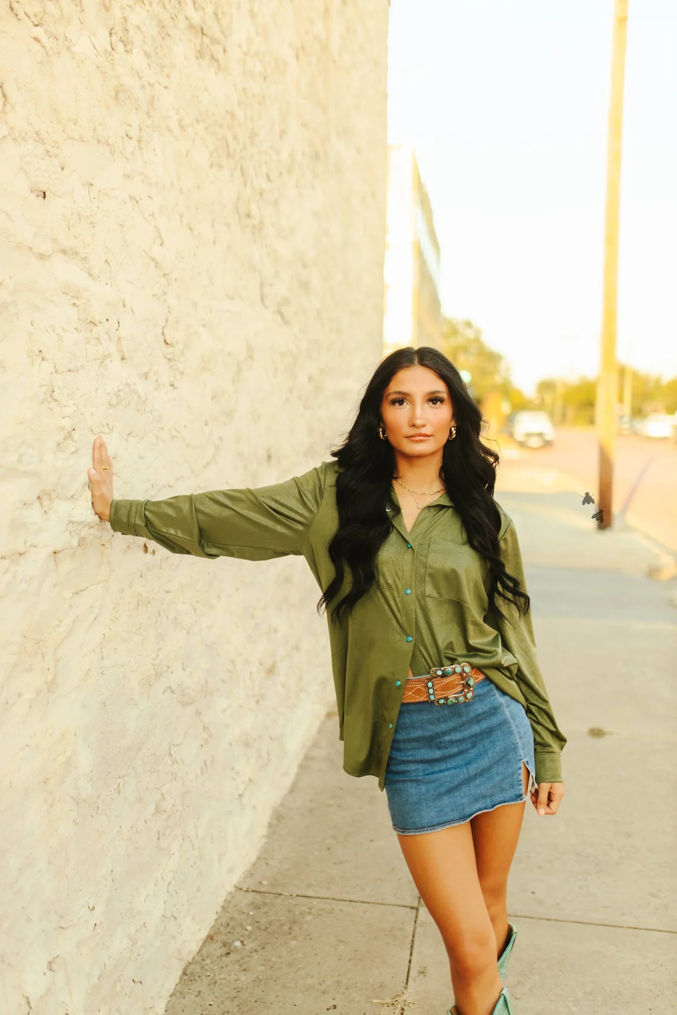 A woman standing with one hand against a wall, wearing a green button-down shirt with turquoise pearl snaps, paired with a blue denim skirt.