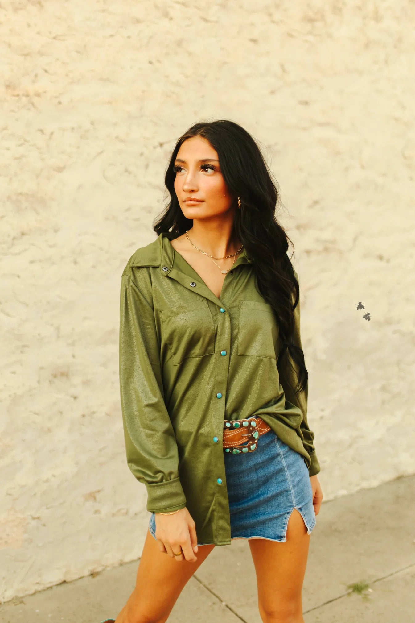 Woman wearing an olive green jacket and denim skirt against a beige wall.