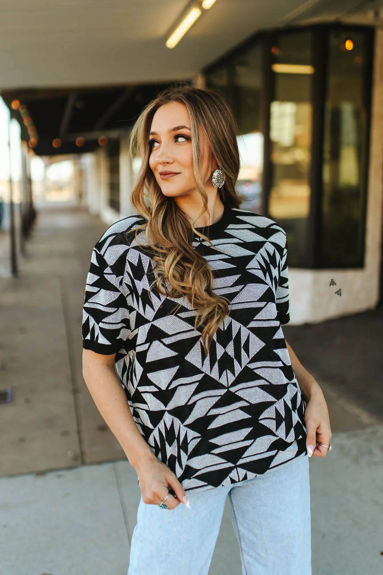 A woman standing outdoors wearing a black and white geometric pattern top with short sleeves, paired with light blue jeans.