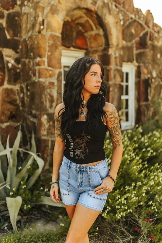 A woman standing outdoors wearing a black sleeveless tank top with lace detailing and blue denim shorts.