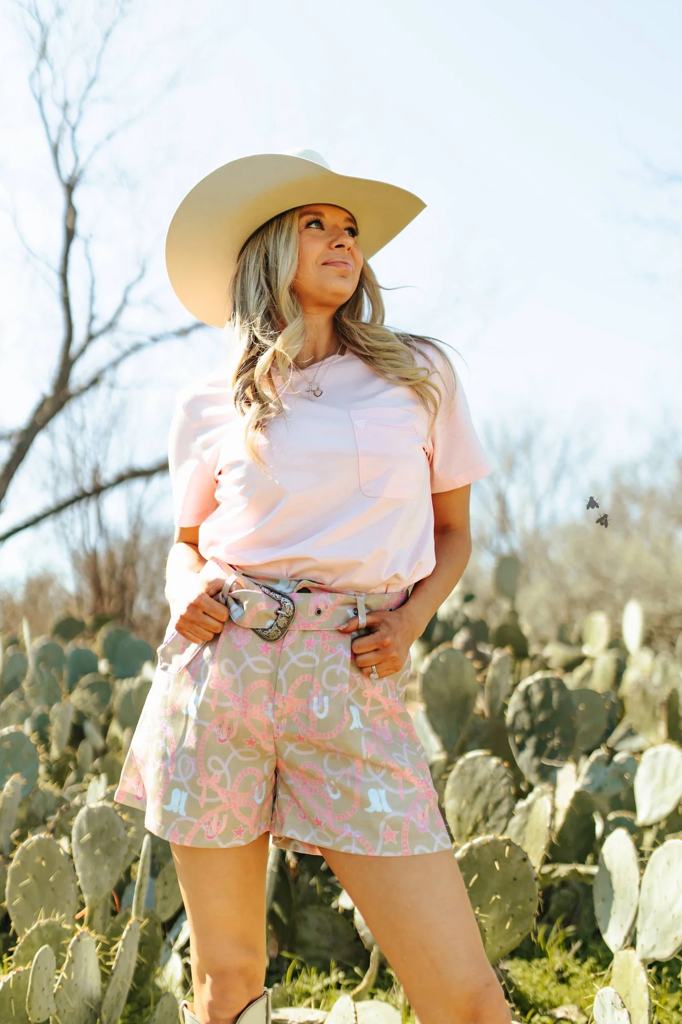 A woman wearing a pink short-sleeved t-shirt and multicolored shorts, with a wide-brimmed hat, standing in a outdoor setting with cacti in the background.