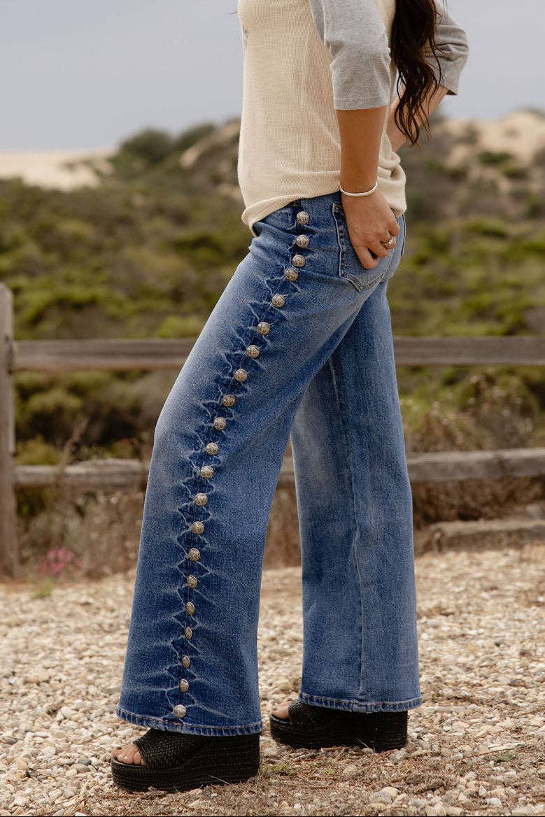 A woman wearing blue wide-leg jeans with decorative gold-tone button details down the side, a cream and gray long-sleeve top, and black platform sandals, standing outdoors on a gravel path with a wooden fence and dunes in the background.
