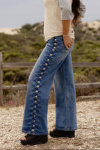 A woman wearing blue wide-leg jeans with decorative gold-tone button details down the side, a cream and gray long-sleeve top, and black platform sandals, standing outdoors on a gravel path with a wooden fence and dunes in the background.