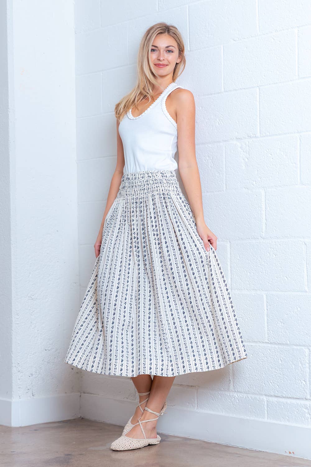 Woman wearing a white sleeveless top and patterned skirt against a white brick wall.
