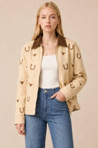A woman modeling a beige jacket with brown horseshoe and longhorn motifs, worn over a white tank top and blue jeans against a solid beige background.
