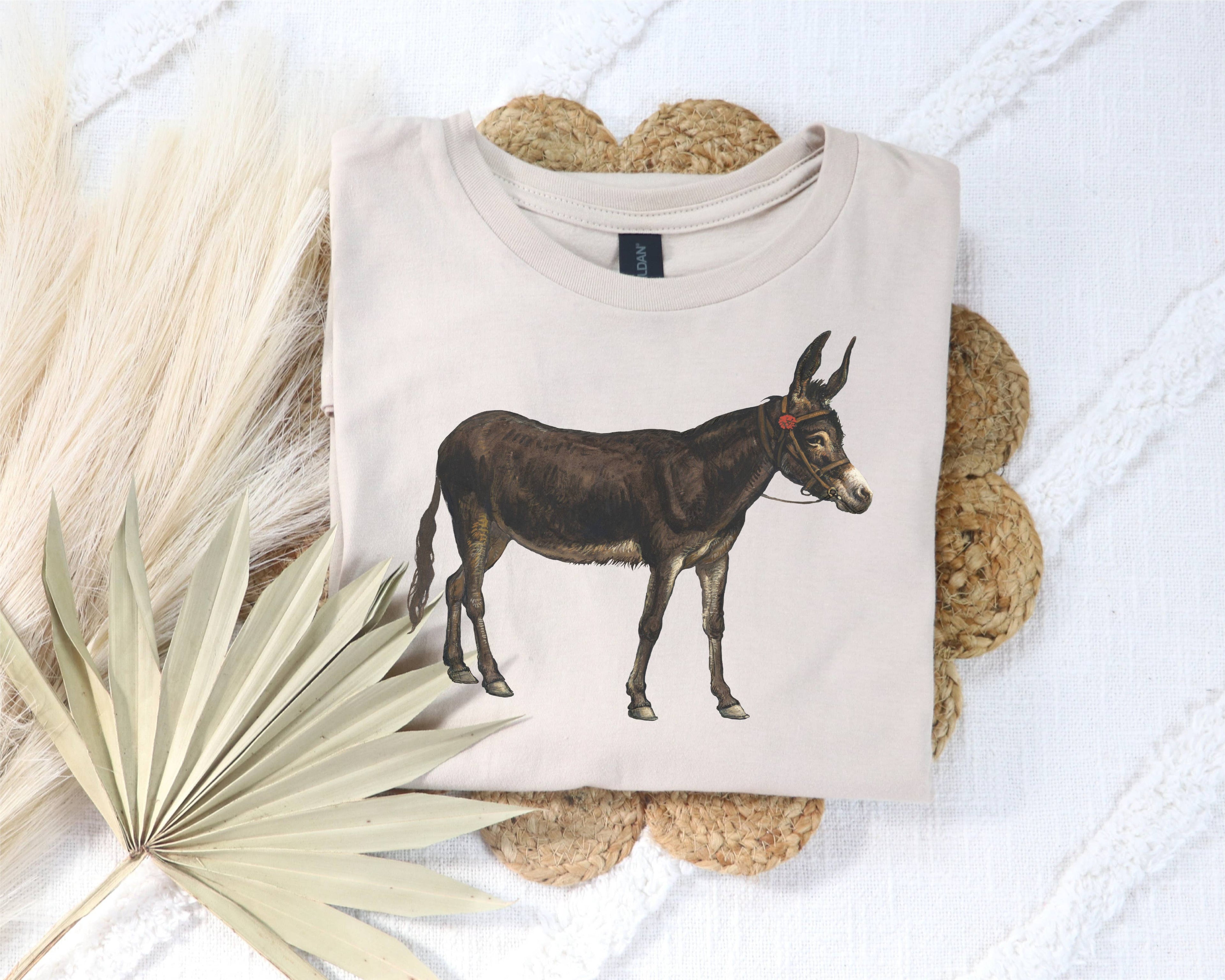 Beige t-shirt with a donkey illustration on a textured surface with dried leaves.