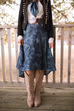 A person wearing a blue patterned midi skirt with a black western-style jacket, white blouse, blue bow, and beige cowboy boots, standing on a wooden deck in front of a railing.