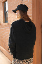 Person wearing a black hoodie and cap, standing against a brick wall.