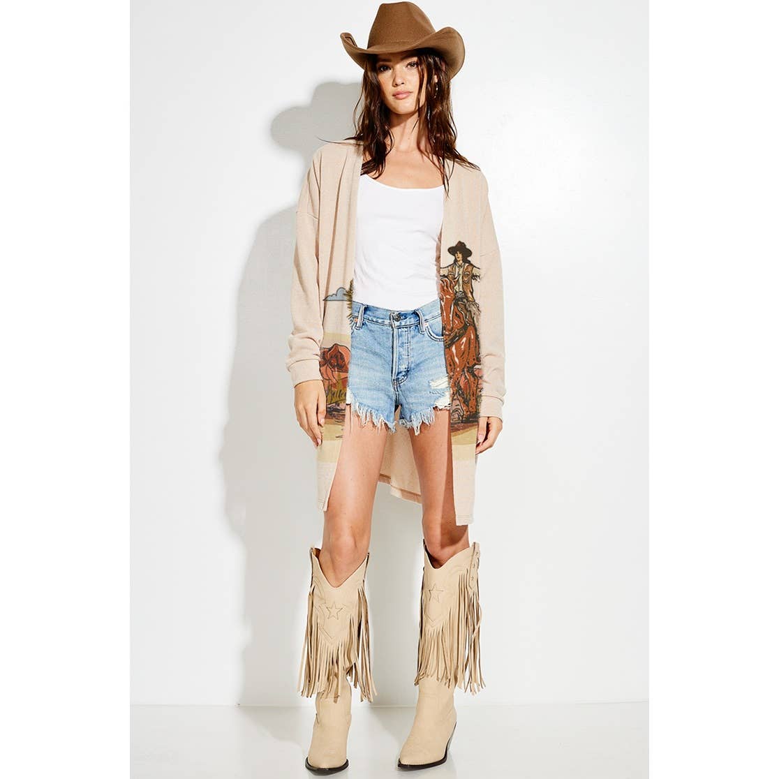 Woman wearing a beige cardigan, white top, denim shorts, and beige fringe boots on a white background