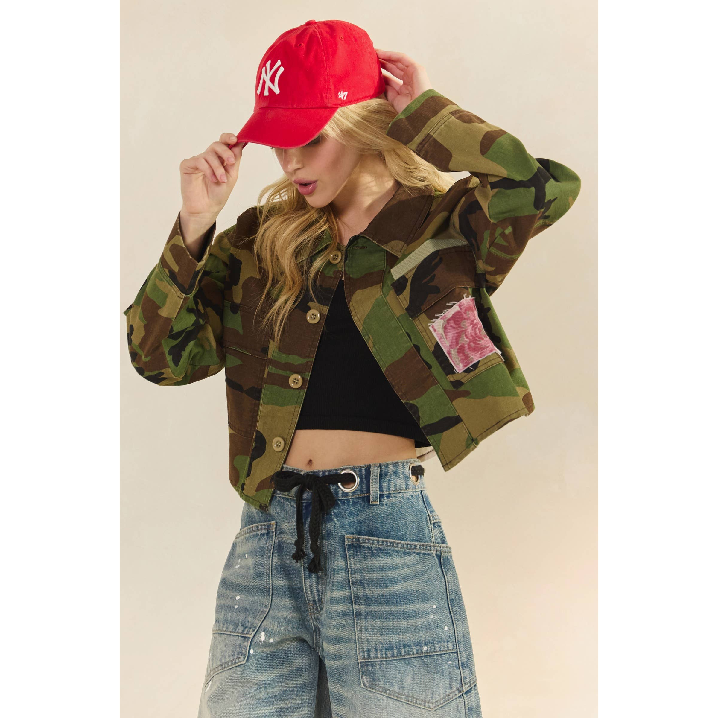 Person wearing a camouflage jacket, black crop top, and blue jeans with a red cap.