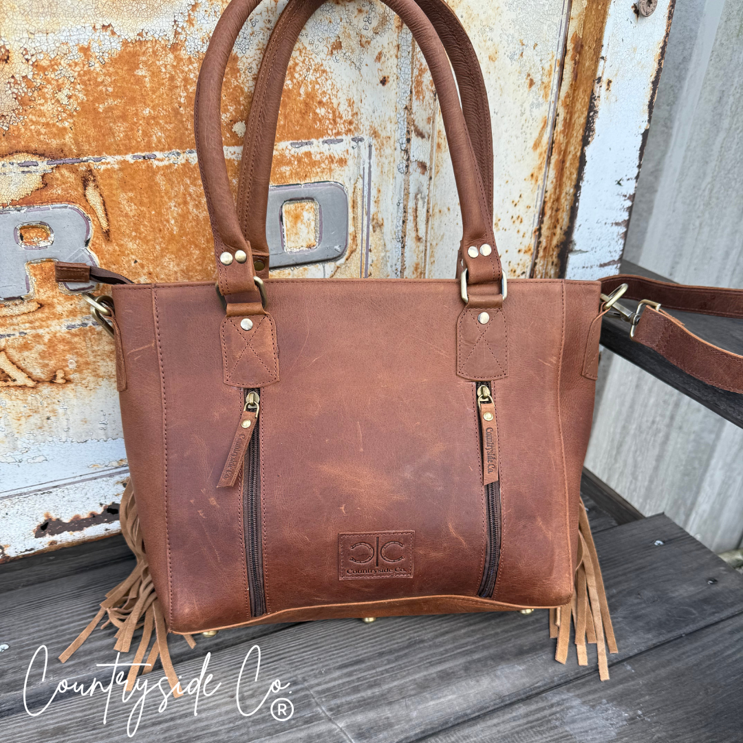 Brown leather handbag with fringe details on a rustic wooden surface, featuring the 'Countryside Co' brand.