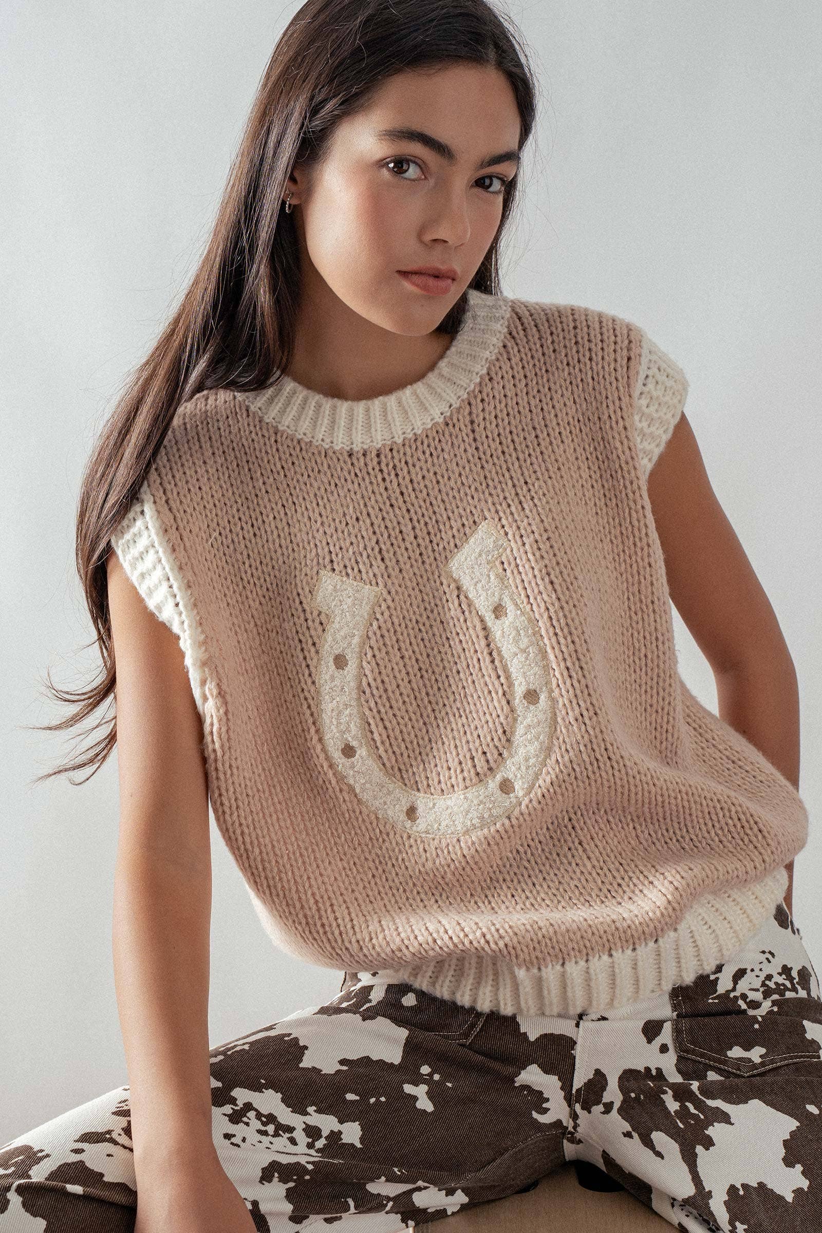 Woman wearing a beige sleeveless sweater with a horseshoe design on a plain background