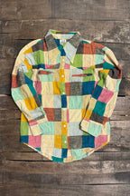 A long-sleeve patchwork button-up shirt made from colorful fabric squares, laid flat on a rustic dark wood plank background.