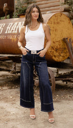 A woman wearing dark blue wide-leg denim jeans with contrast stitching, a white tank top, and beige heeled sandals, standing outdoors in front of a large rusted yellow-brown tanker truck.