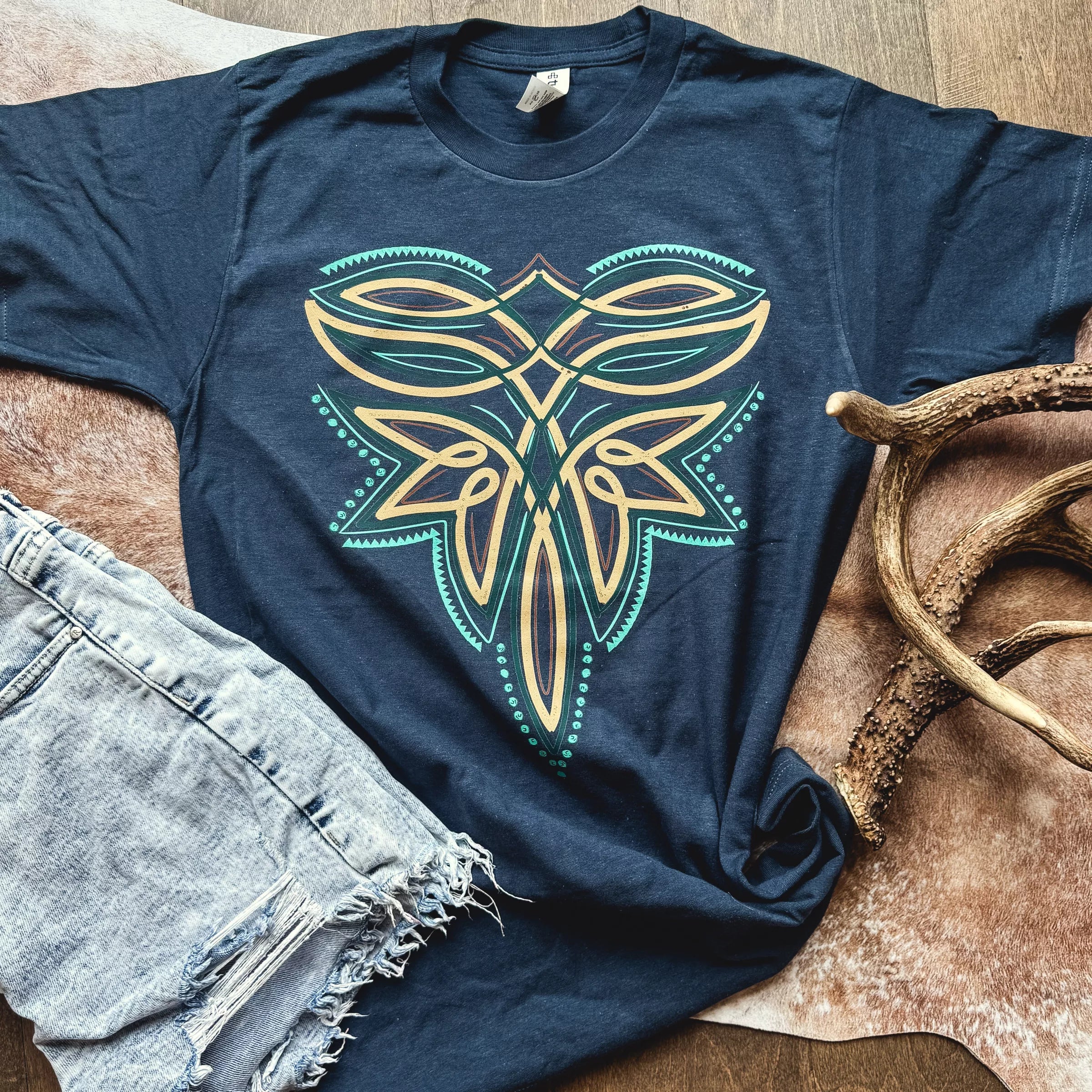 Navy blue t-shirt with a geometric deer antler design, denim shorts, and antlers on a wooden surface.