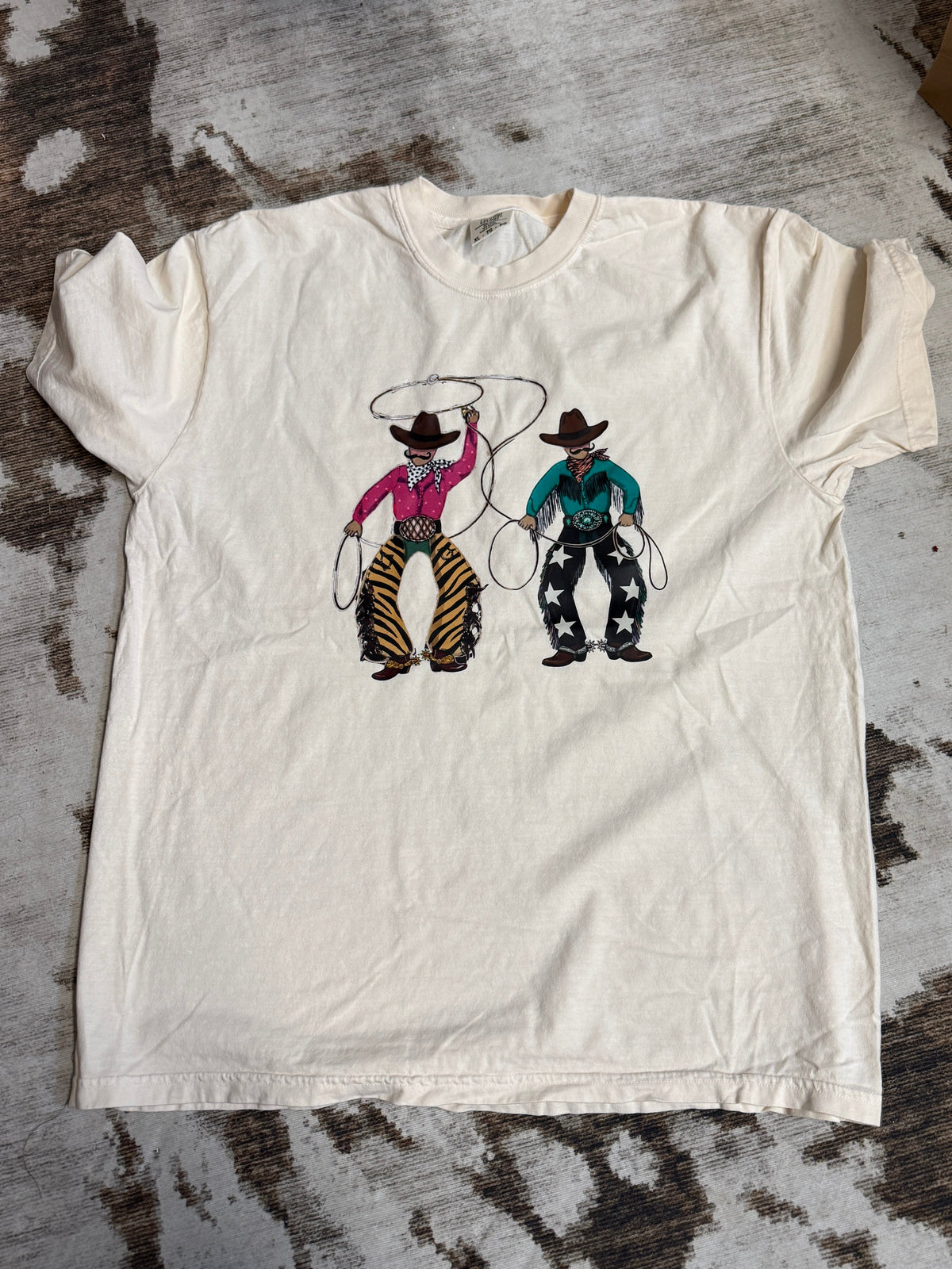 An off-white short-sleeve t-shirt laid flat on a mottled gray and brown floor, featuring a graphic of two western cowboys with lasso designs.