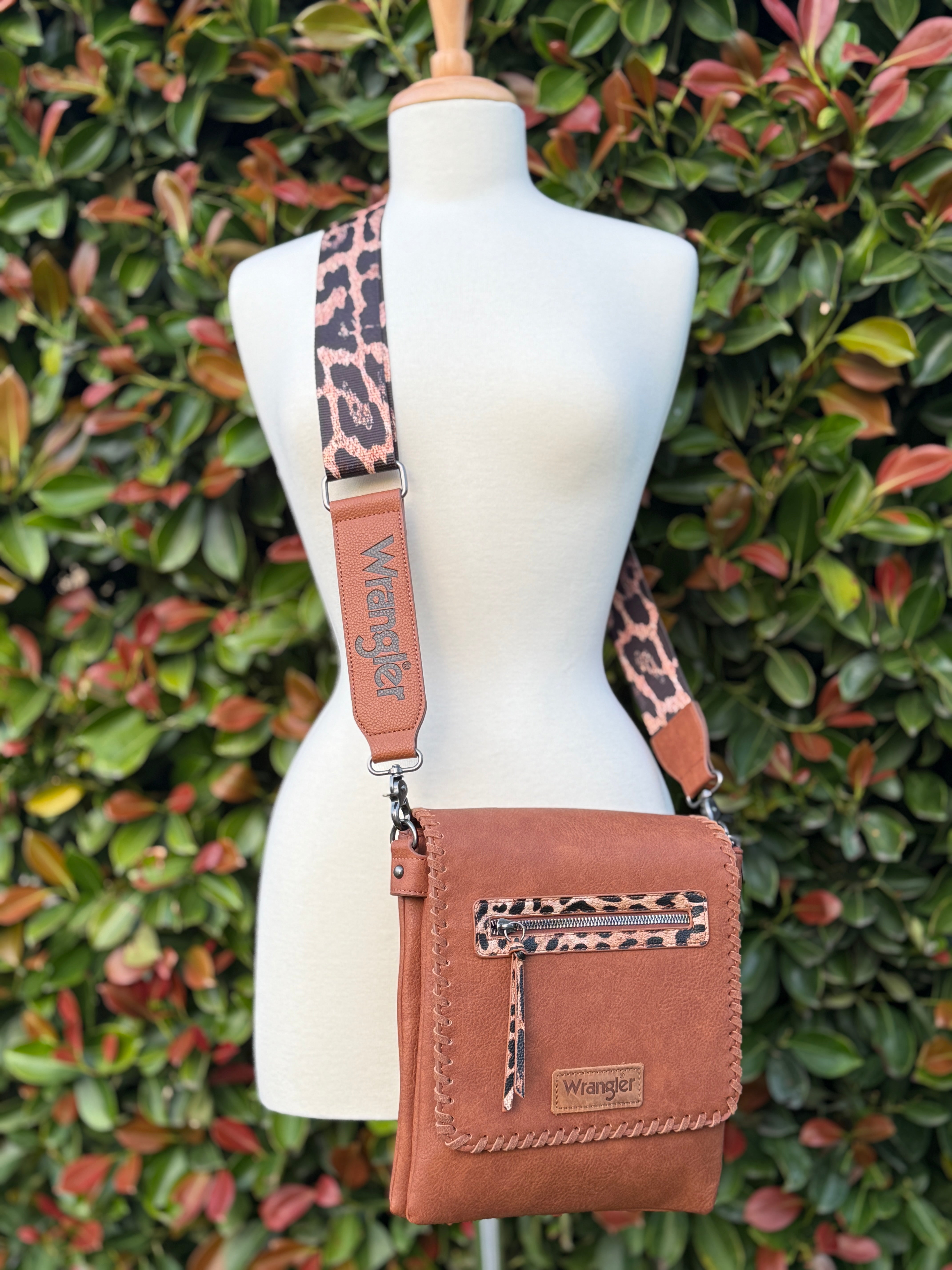 A crossbody bag with dark brown PU leather, detailed flap with magnetic closure, front zip pocket, and an adjustable leopard-print strap, displayed on a mannequin against a leafy background.