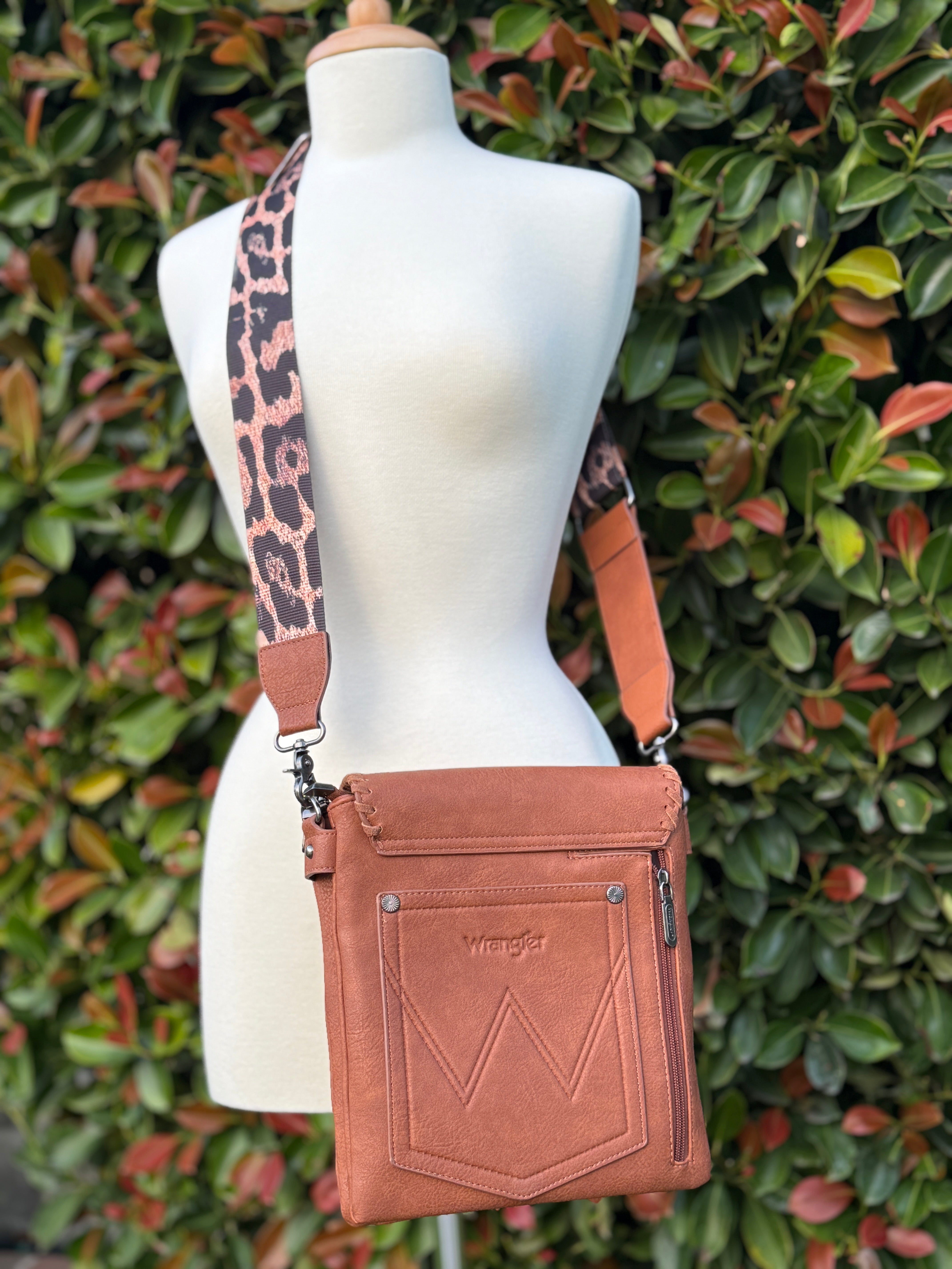 A crossbody bag with dark brown PU leather, detailed flap with magnetic closure, front zip pocket, and an adjustable leopard-print strap, displayed on a mannequin against a leafy background.