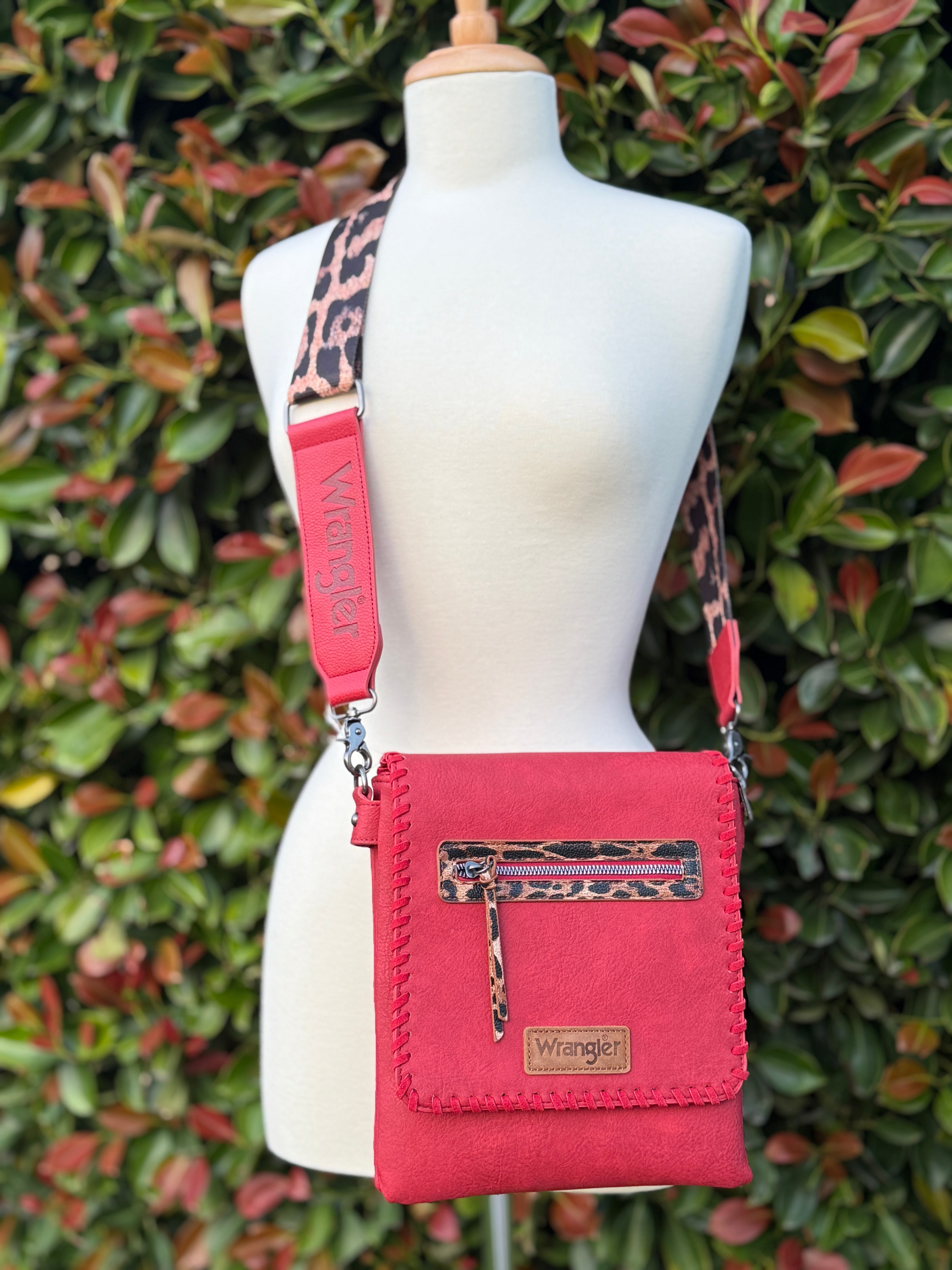 A red crossbody bag with whipstitch detailing and a leopard print adjustable strap, displayed on a mannequin against a leafy background.