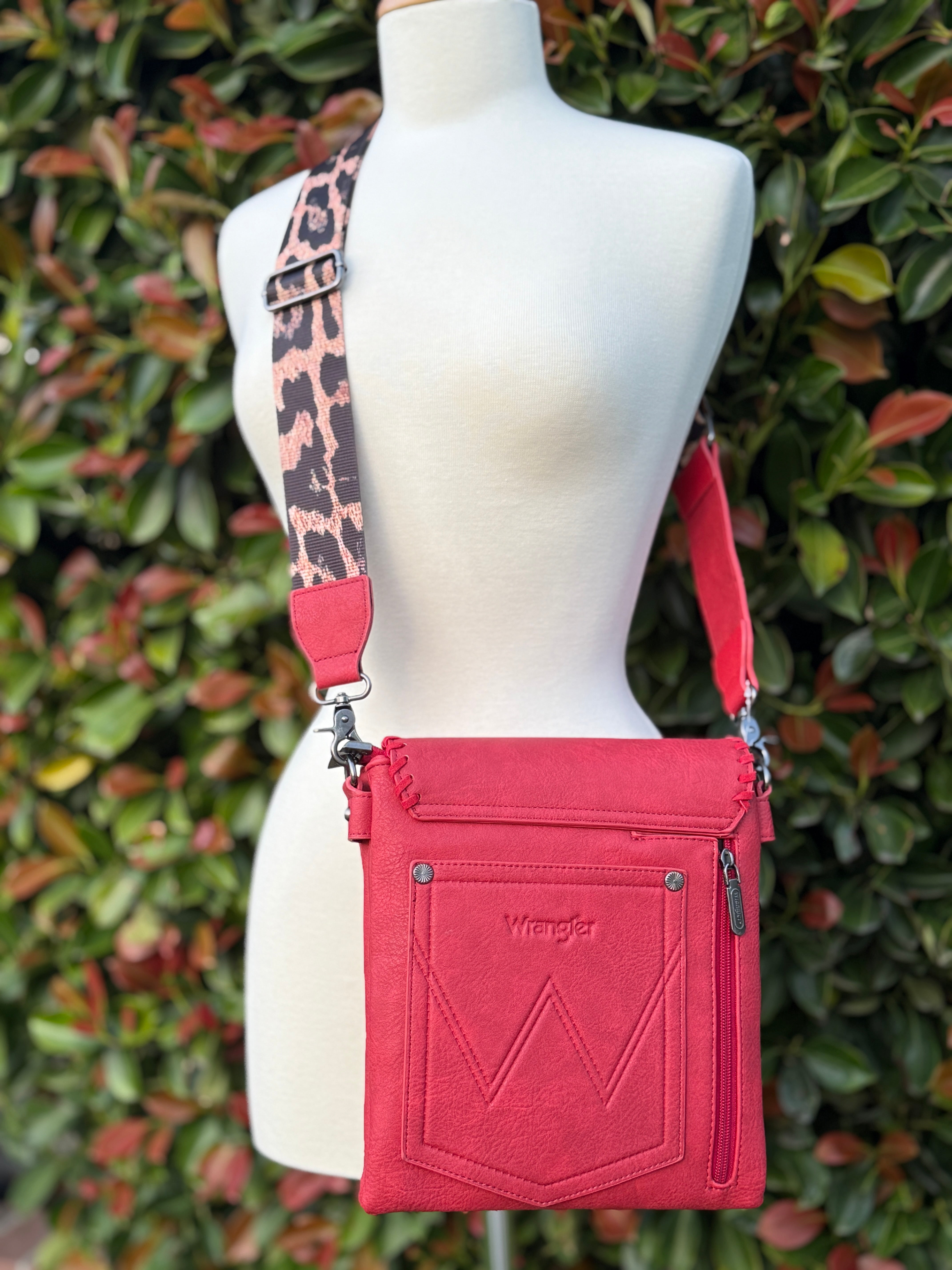 A red crossbody bag with whipstitch detailing and a leopard print adjustable strap, displayed on a mannequin against a leafy background.