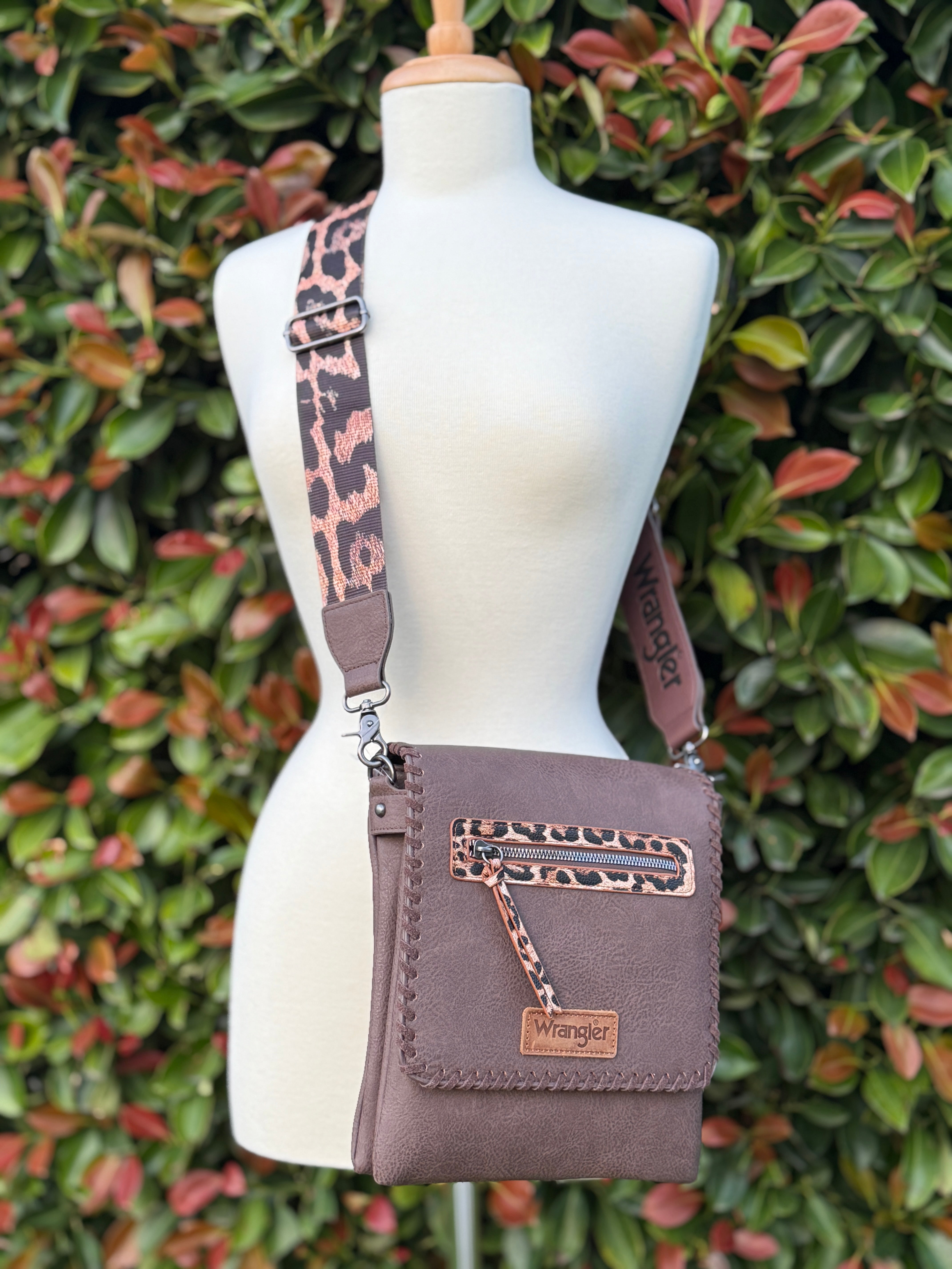 A brown crossbody bag with leopard print strap displayed on a mannequin against a leafy background.