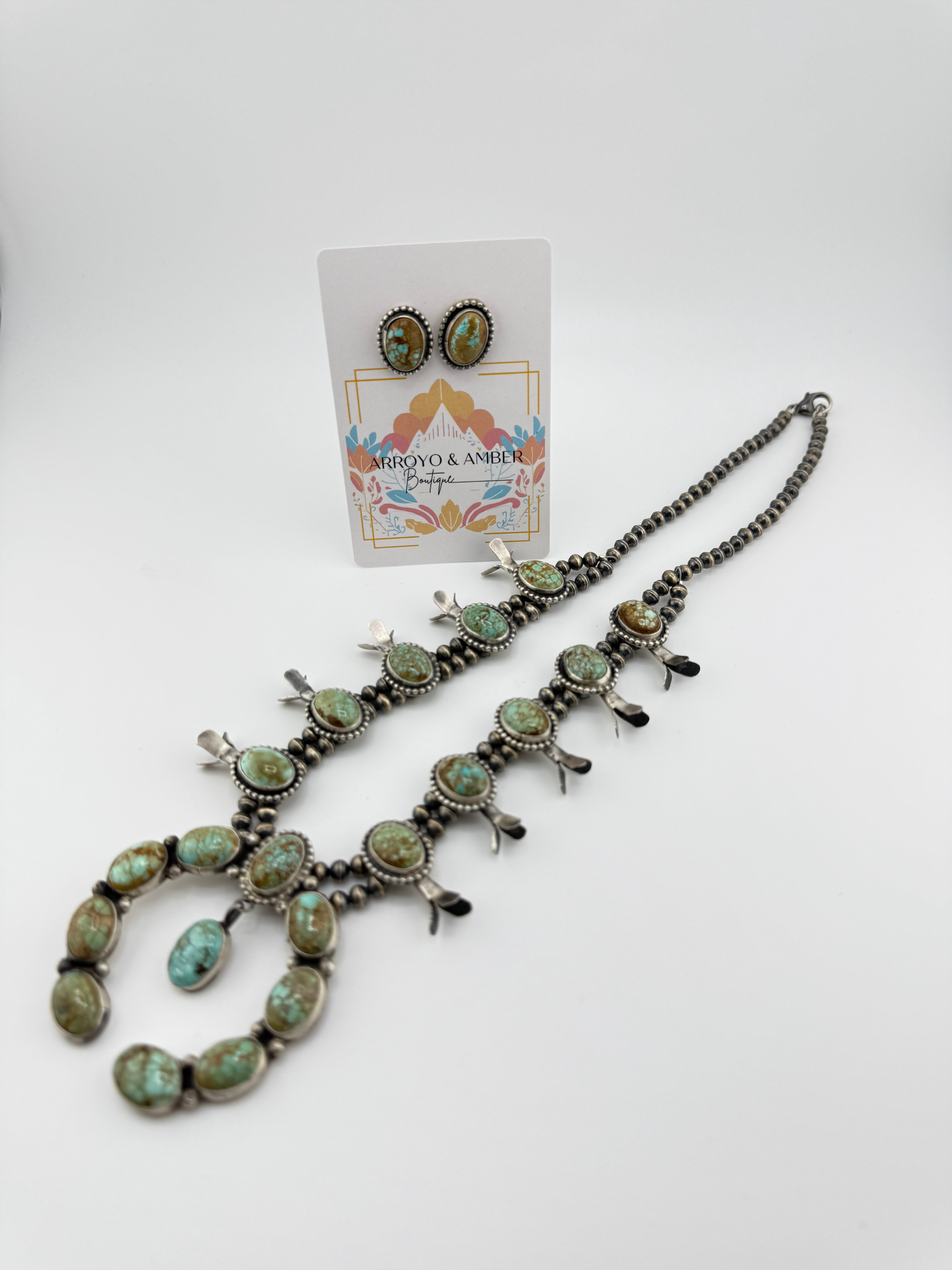 Necklace with turquoise stones and silver accents on a white background, with a card displaying brand name.