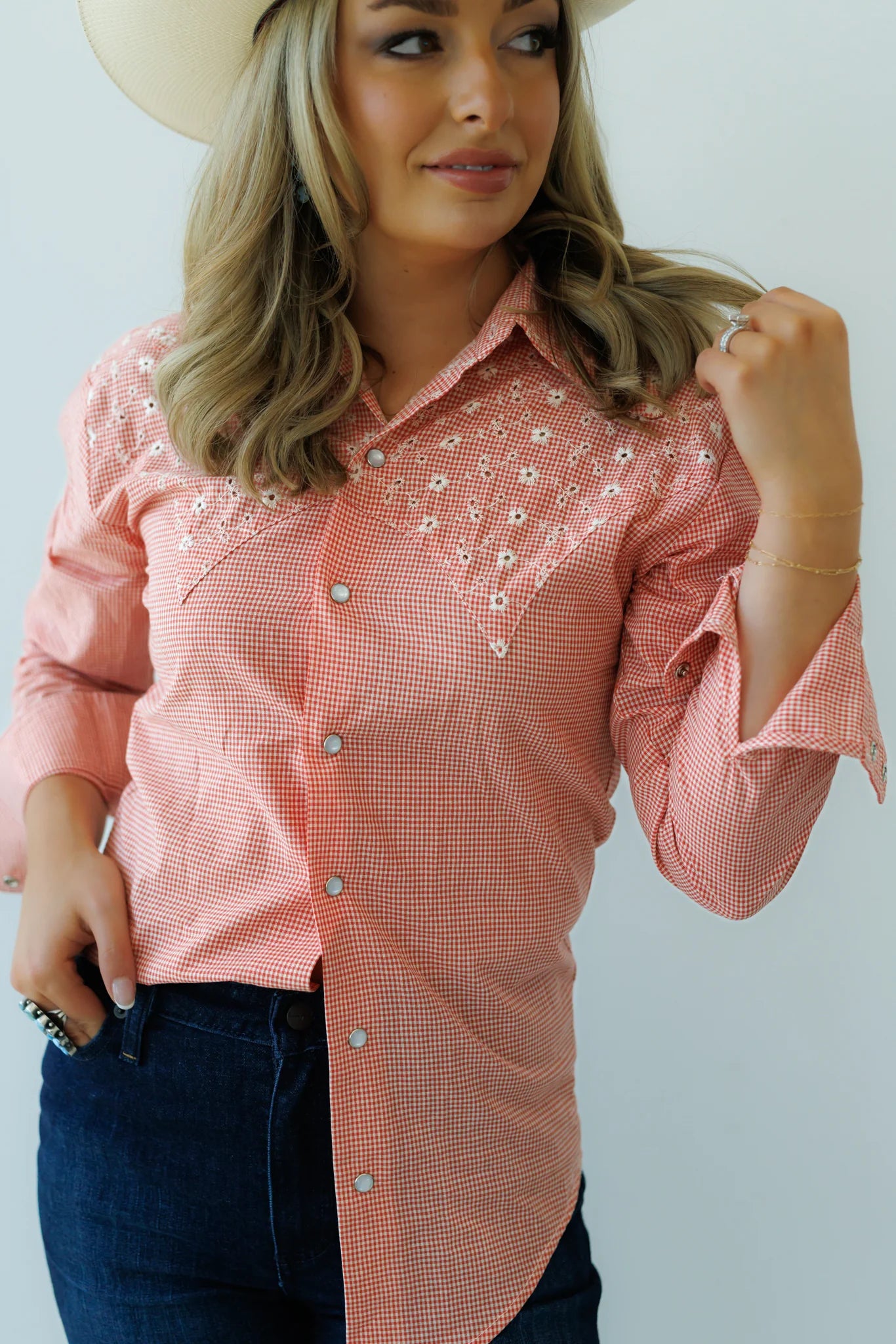 A woman wearing a pink and white gingham western-style button-down shirt with floral embroidery on the chest, paired with dark blue jeans and a cream cowboy hat.