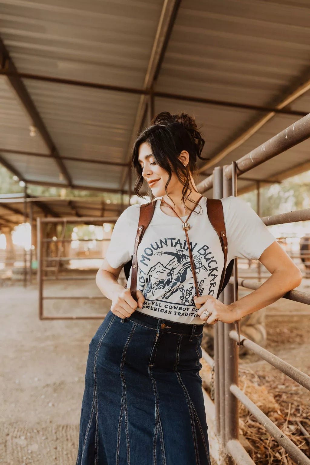 Woman wearing a graphic t-shirt and overalls standing in an outdoor setting with a rustic background.