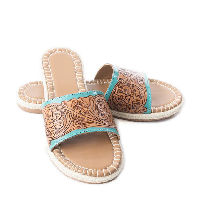 A pair of tooled leather slides with turquoise accents and floral patterns, displayed against a white background.