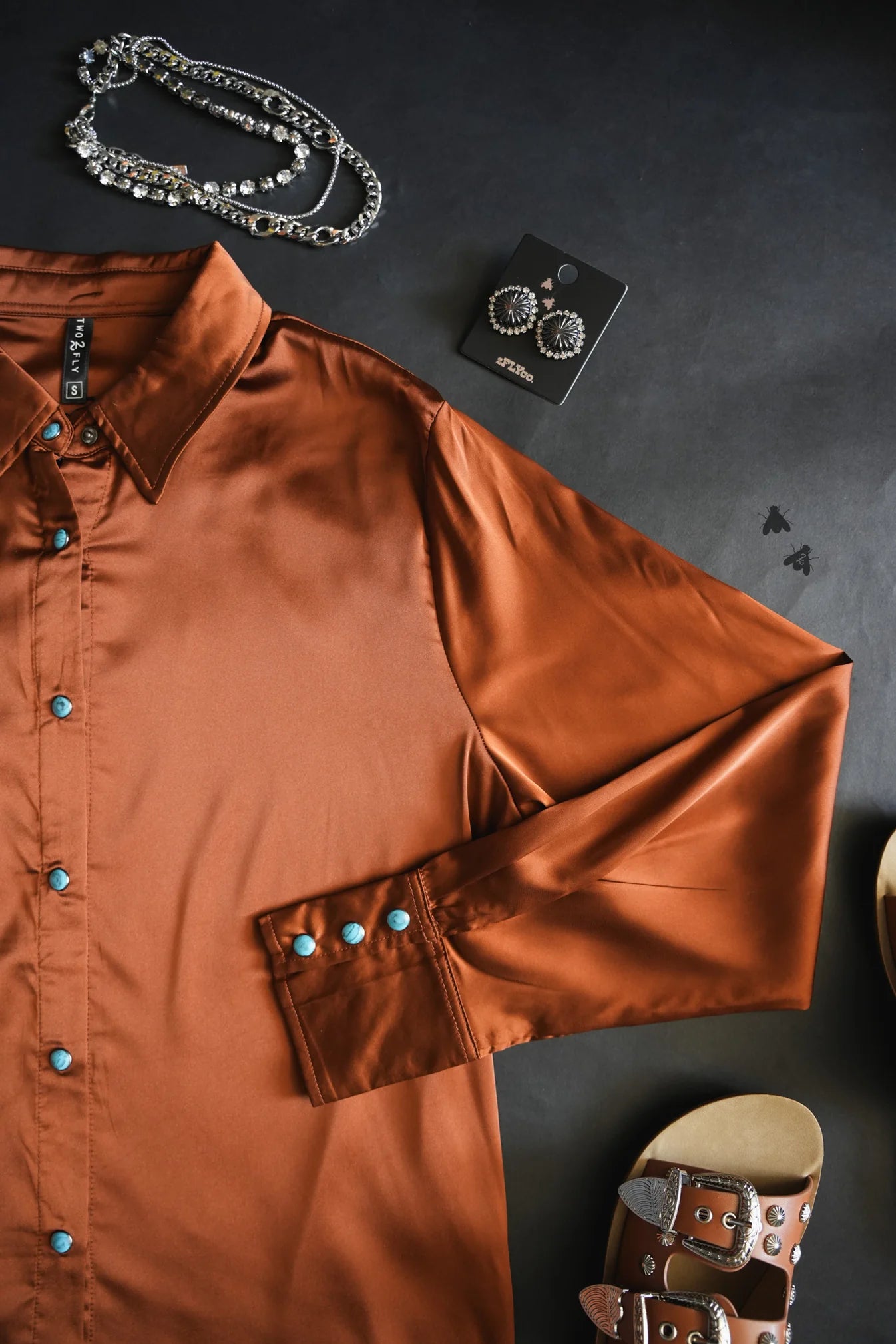 Brown shirt with turquoise buttons on a dark surface with jewelry and a sandal.