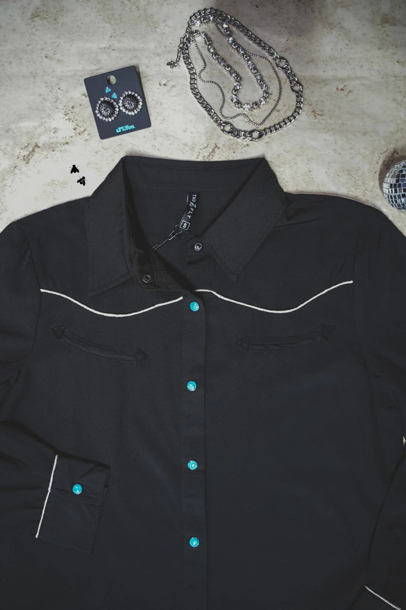 Black shirt with turquoise buttons and white piping on a textured surface.