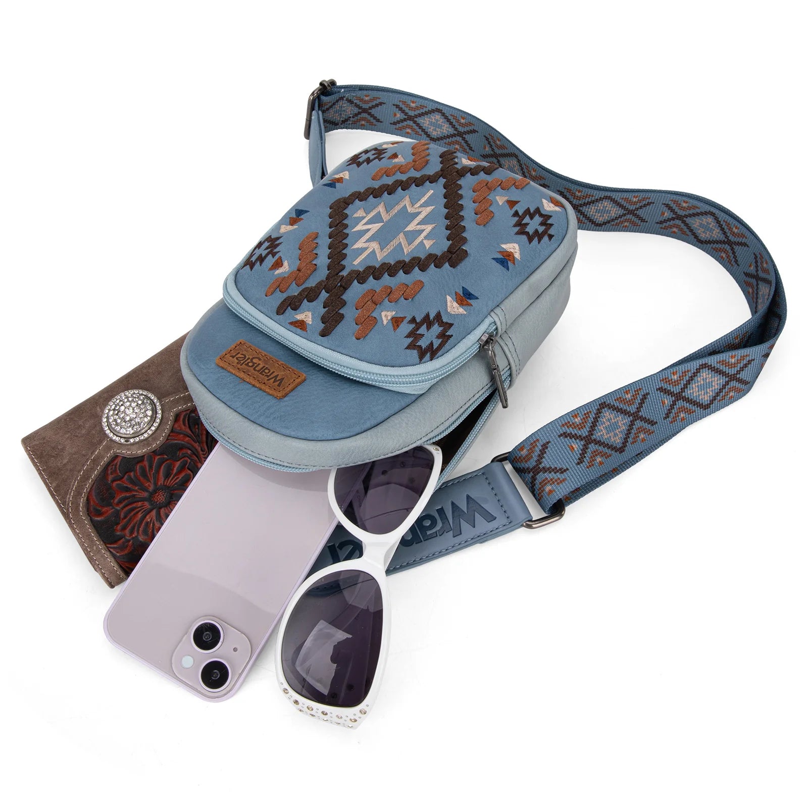 A blue Wrangler Aztec Embroidered Sling Bag displayed on a mannequin against a foliage background.