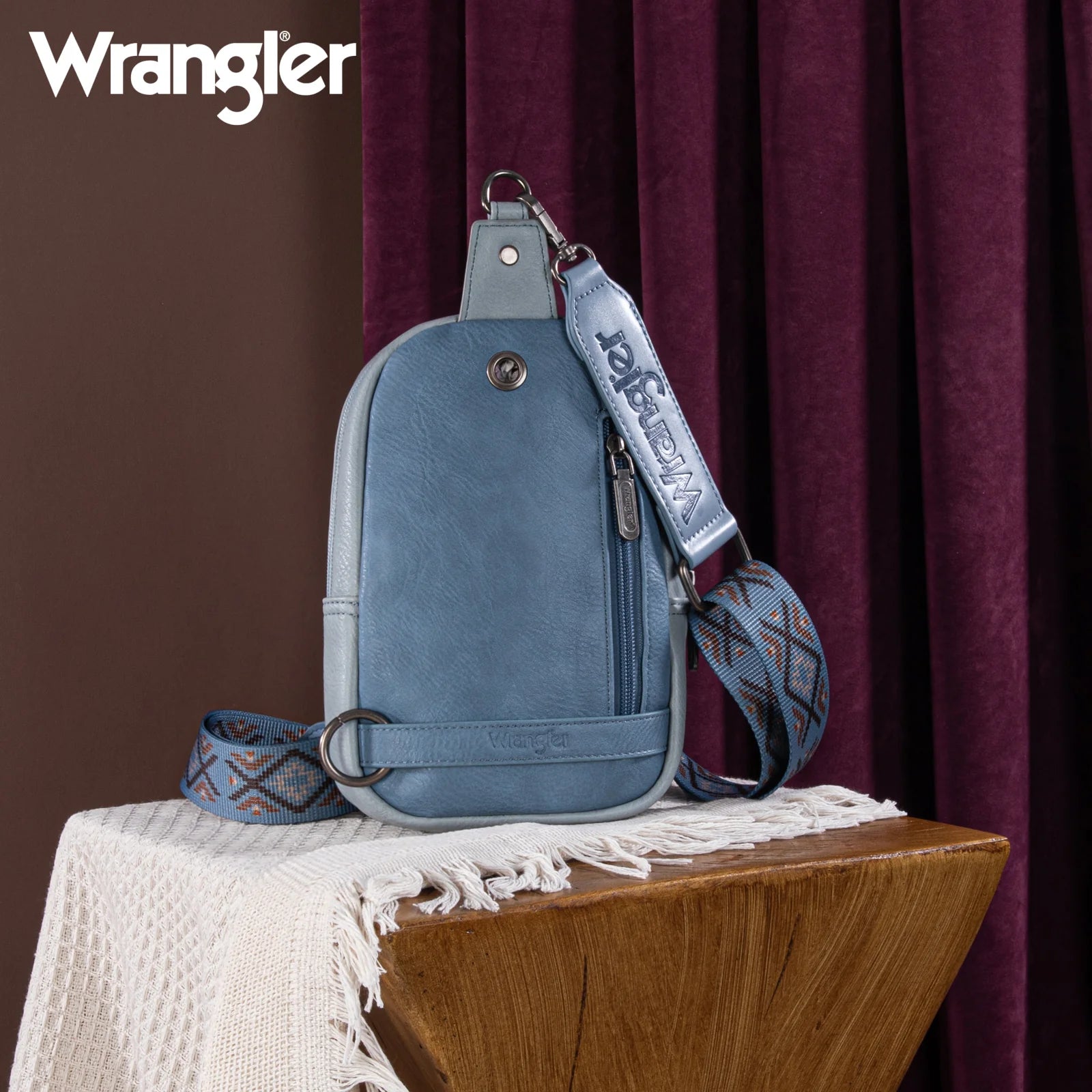 A blue Wrangler Aztec Embroidered Sling Bag displayed on a mannequin against a foliage background.