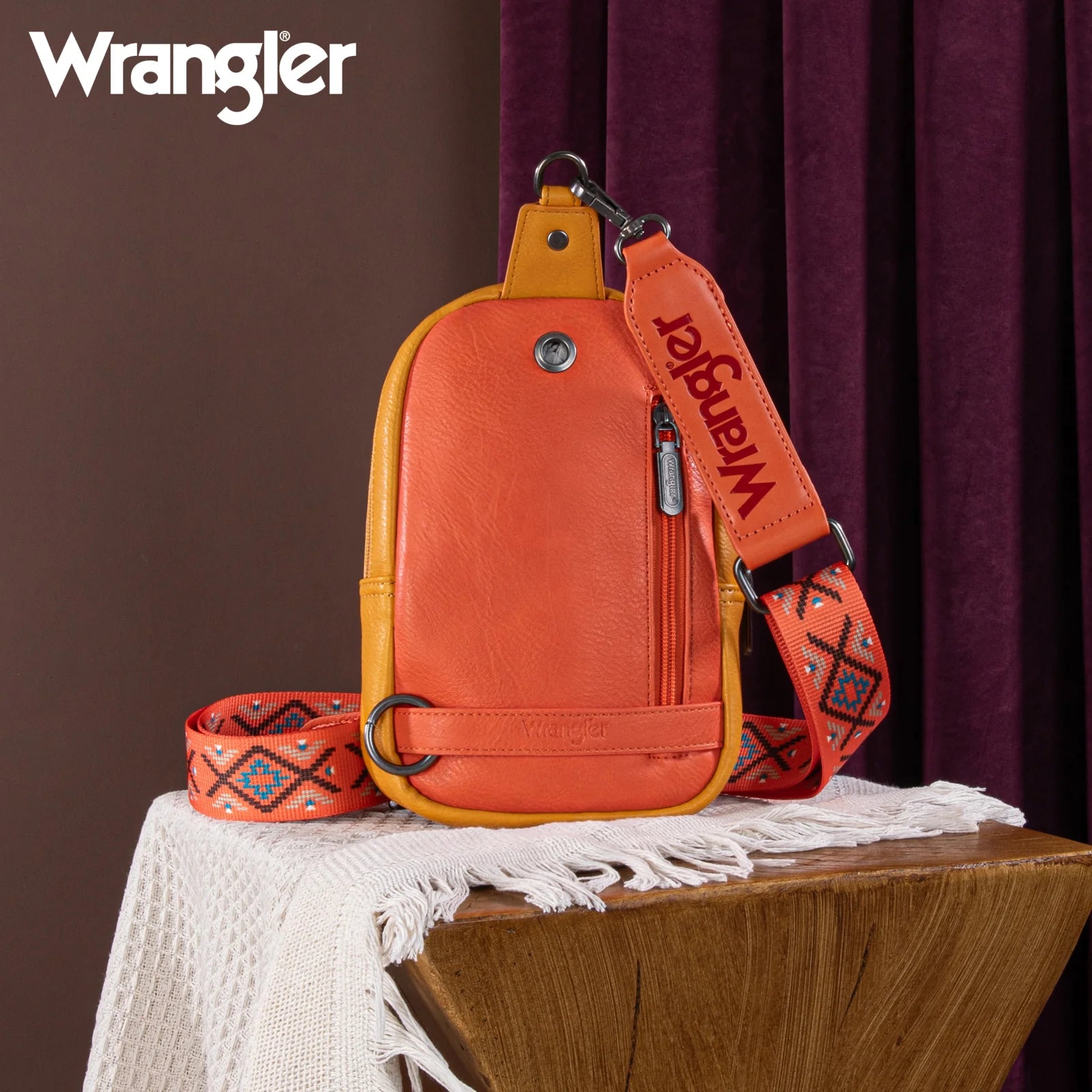 A bright orange and multicolor geometric embroidered Wrangler Southwestern sling bag displayed on a mannequin against a leafy background.