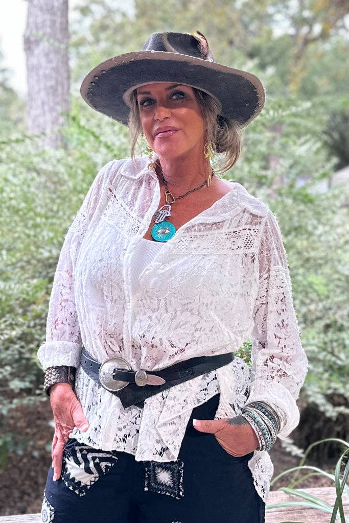 Woman wearing a white lace shirt, black hat, and black pants with a belt outdoors.