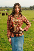 A woman stands in a grassy field wearing a rust-orange plaid button-up shirt over a graphic tee, a long blue denim skirt, and a brown belt with a silver concho buckle. Cows are visible in the background.