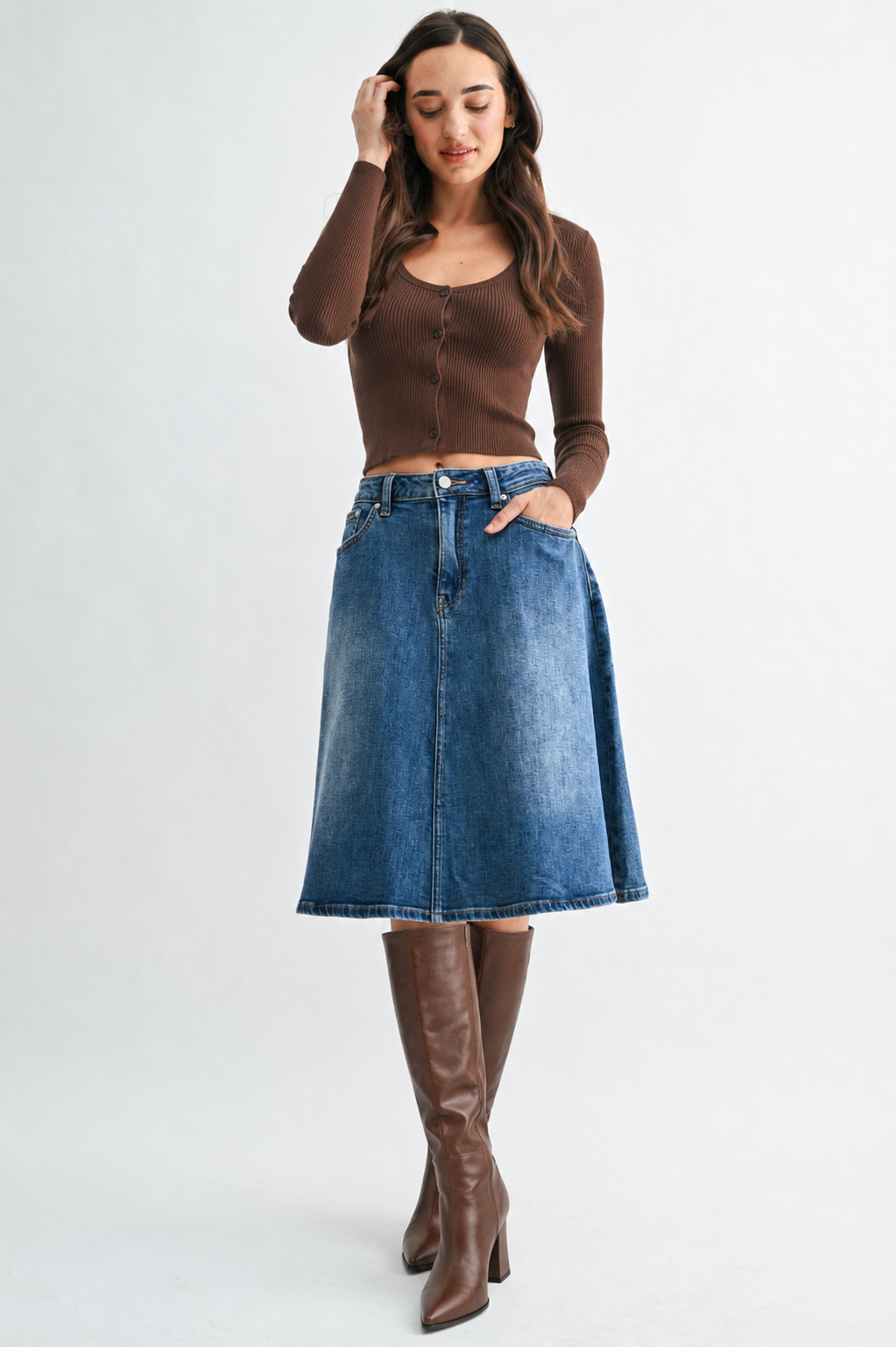 A woman modeling a brown long-sleeve ribbed knit top and a blue denim A-line skirt, paired with brown knee-high leather boots, against a light gray studio background.