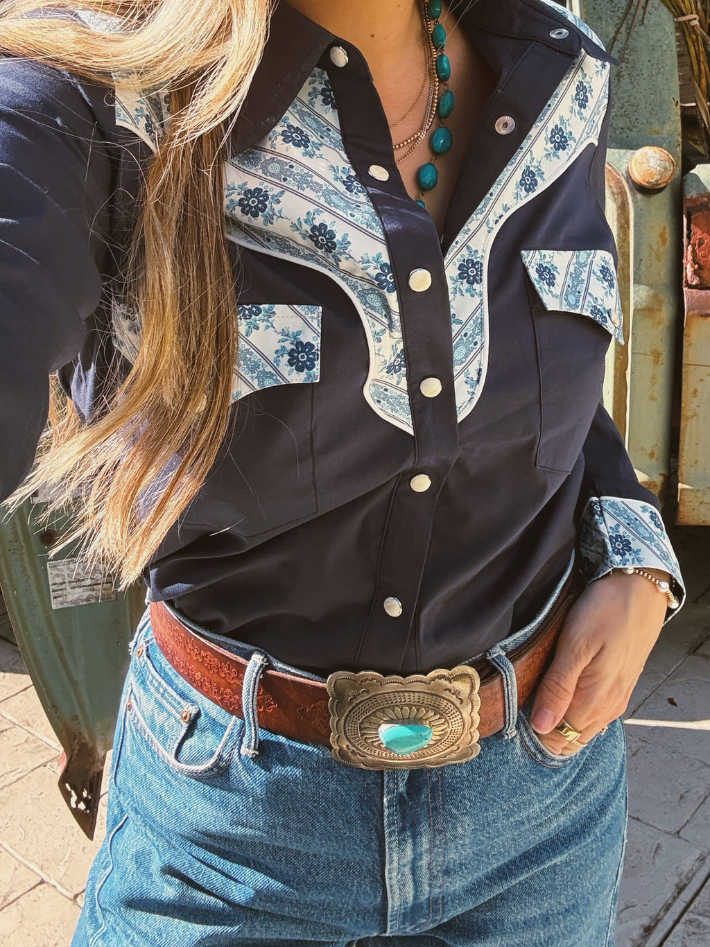 Person wearing a black and white patterned shirt with blue jeans and a turquoise belt buckle.