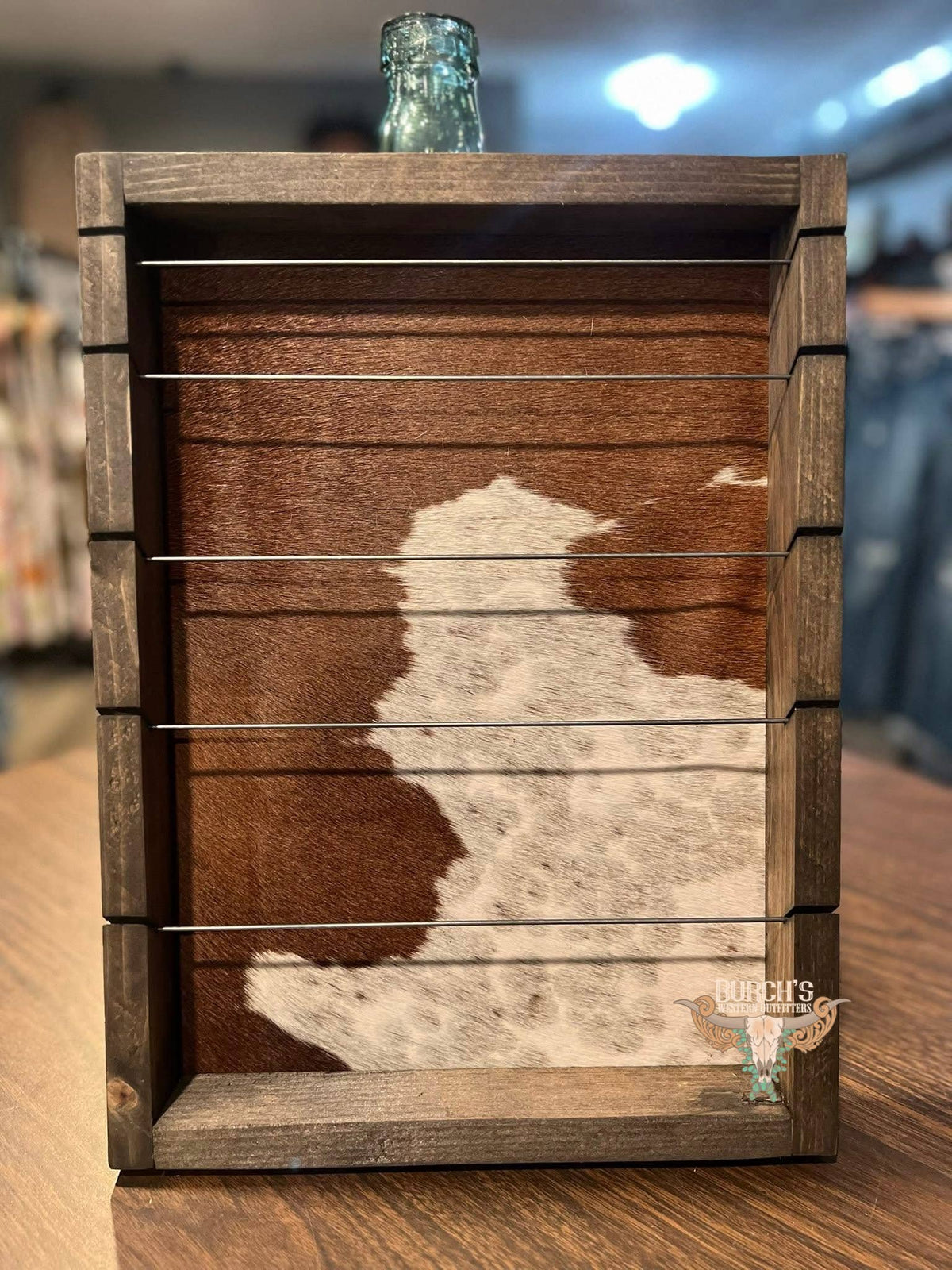 A rustic wooden slat rack or shelf with a cowhide-patterned backing, shown on a wooden table in a store setting with a glass bottle resting on top.