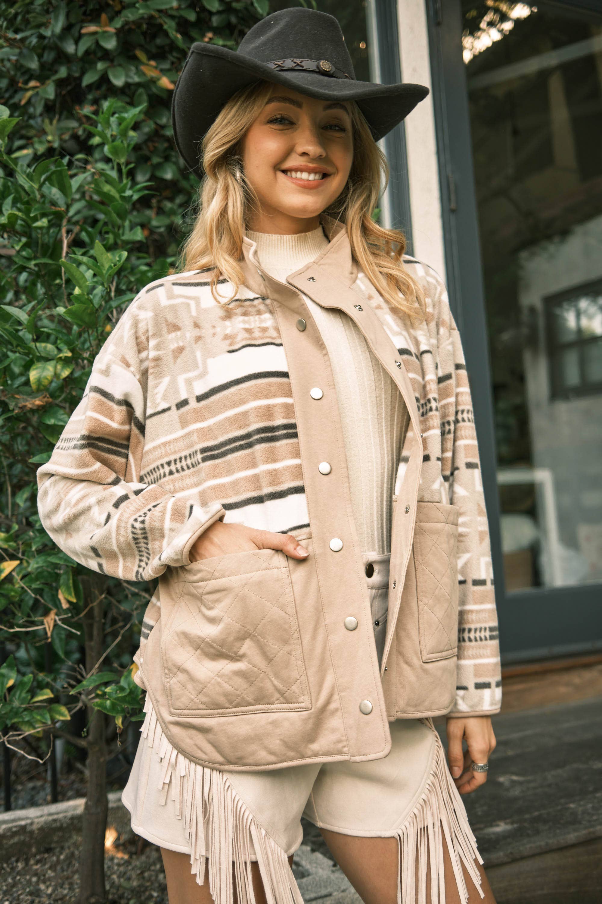 Woman wearing a patterned jacket with fringe, white shirt, and shorts outdoors.