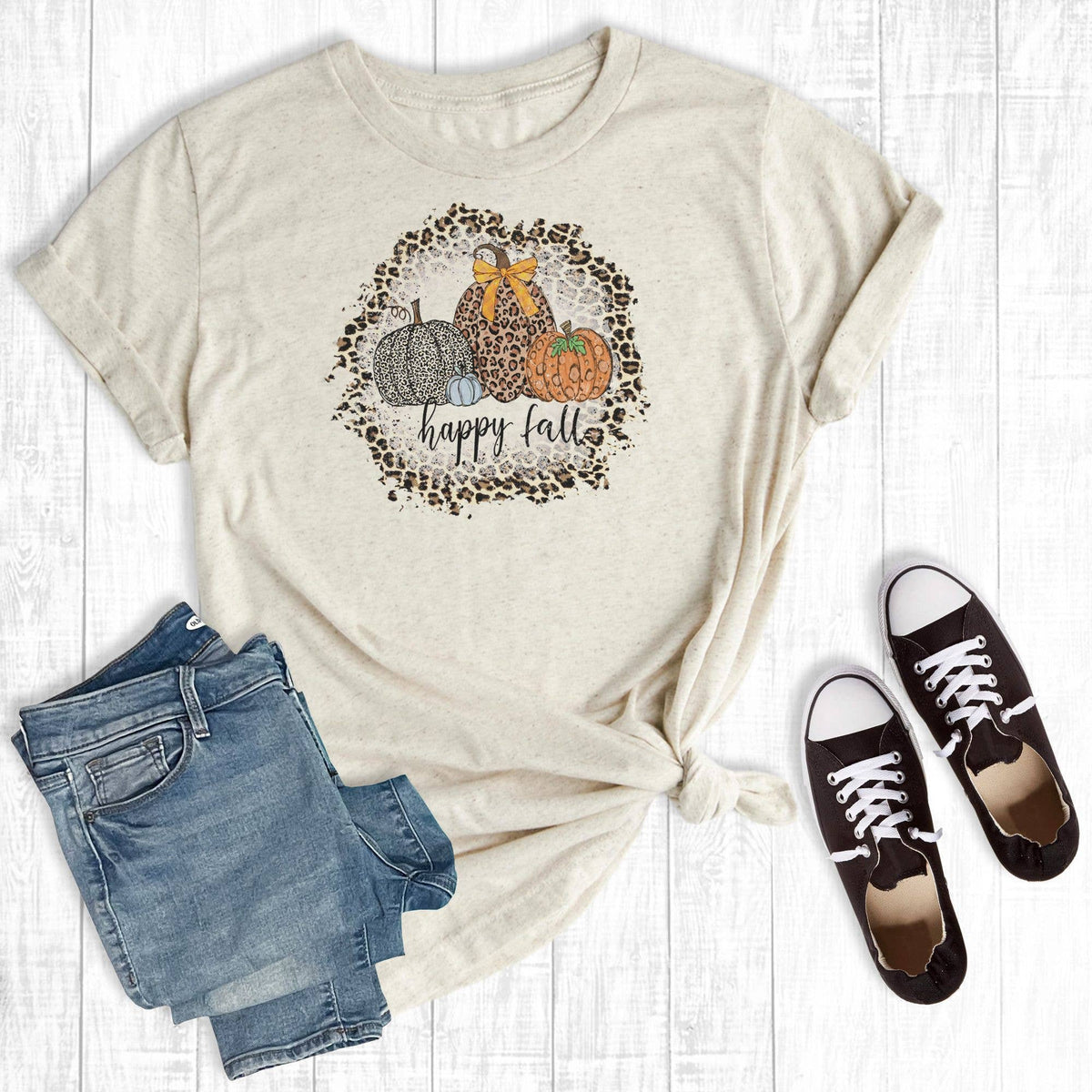 A beige graphic T-shirt laid flat on a white wood background, styled with folded blue jeans and black low-top sneakers. The shirt has a 'happy fall' autumn graphic with pumpkins inside a leopard print circle.