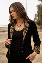 A woman wearing a black velvet blazer with leopard print trim, styled with a black top and a gold pendant necklace, photographed outdoors.