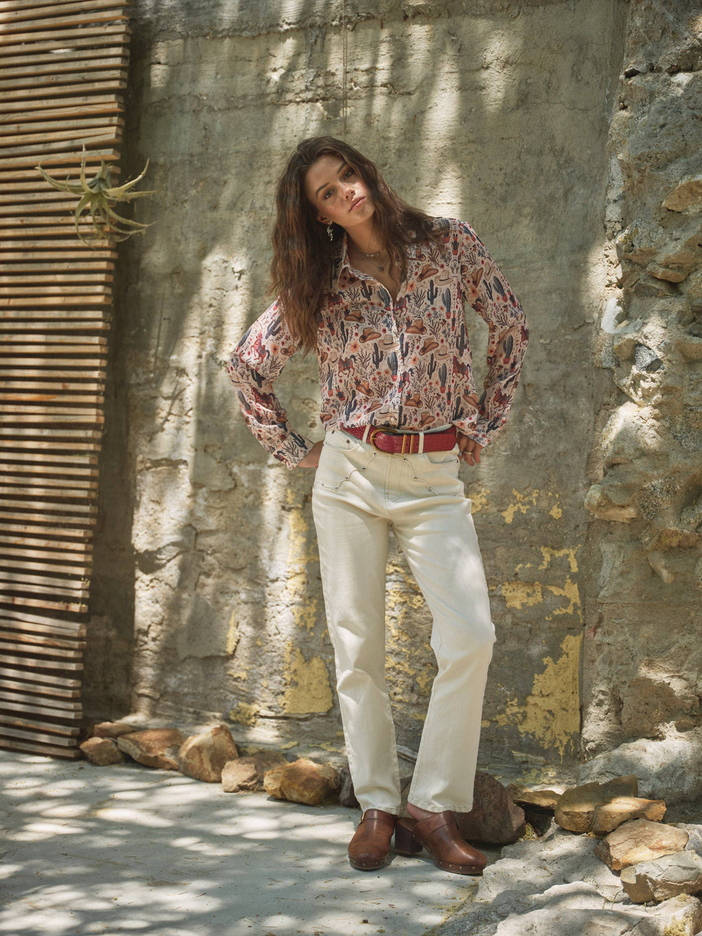 Woman in a patterned shirt and white pants standing against a stone wall.