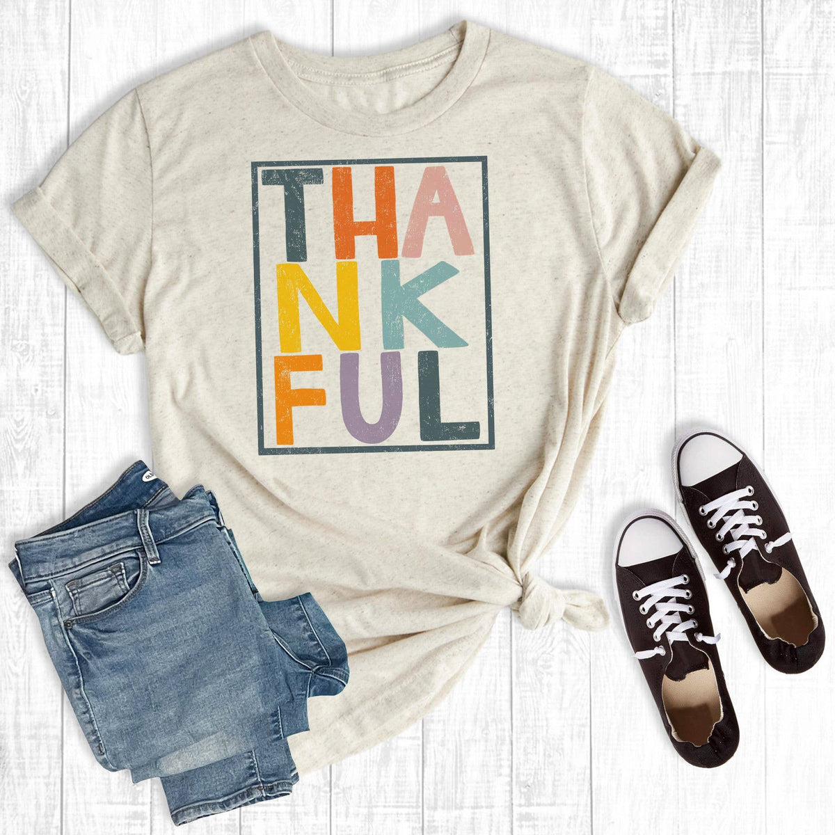 A styled flat-lay of a beige graphic T-shirt with the word 'THANKFUL' printed in large multicolored letters, paired with blue jeans and black low-top sneakers on a white wood plank background.