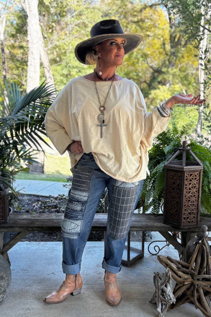 Woman in a beige sweater, plaid pants, and cowboy boots standing outdoors with plants and a bench in the background.