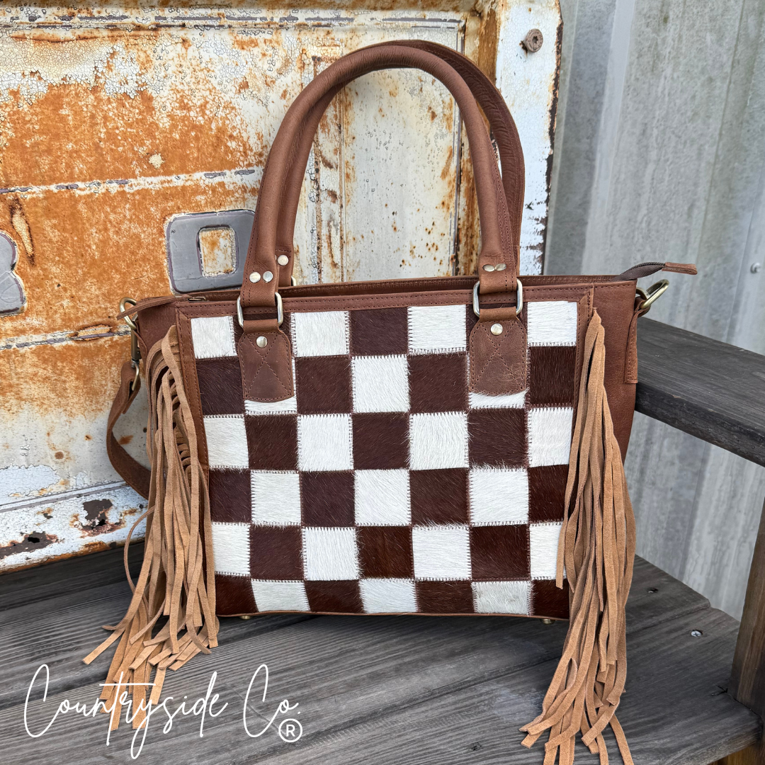 Brown and white checkered handbag with fringes on a rustic wooden surface, featuring 'Countryside Co' branding.