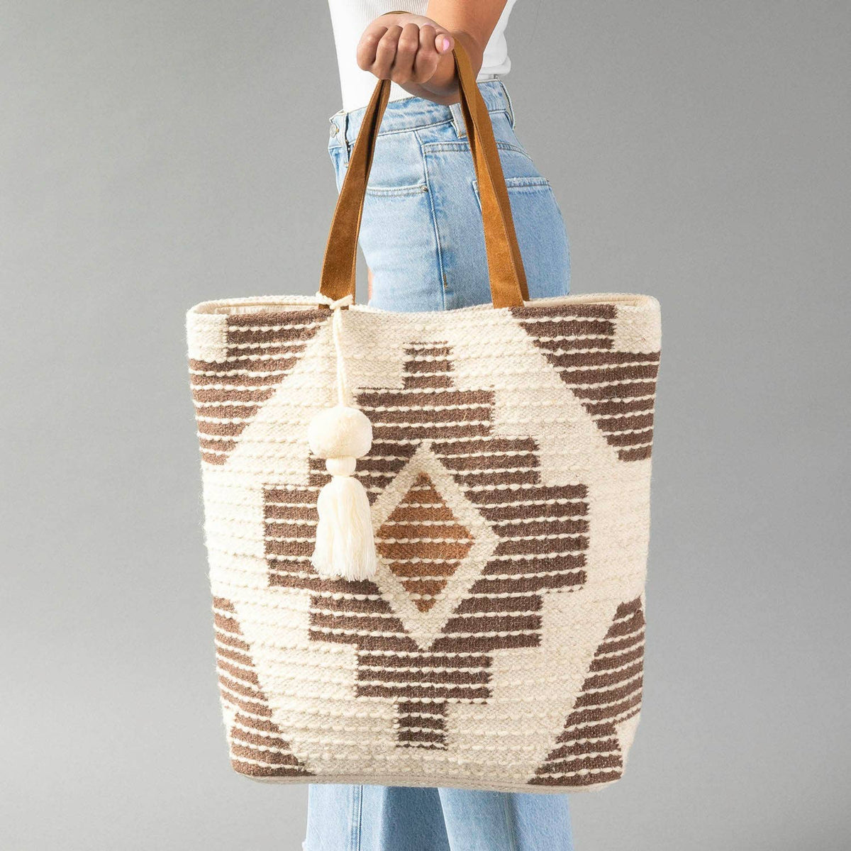 A woven tote bag with a brown and cream geometric pattern, held by a person wearing light blue jeans against a solid gray studio background.